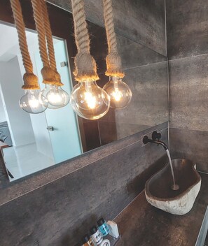 A bathroom with a large mirror, a stone sink, and three hanging light bulbs. - Finikas Suites & Apartments Airport (Spata-Artemida)