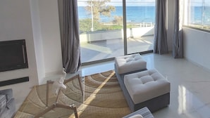 A modern living room with a large window offering a view of the sea, a sofa, a coffee table, and a hanging light fixture. - Finikas Suites & Apartments Airport (Spata-Artemida)
