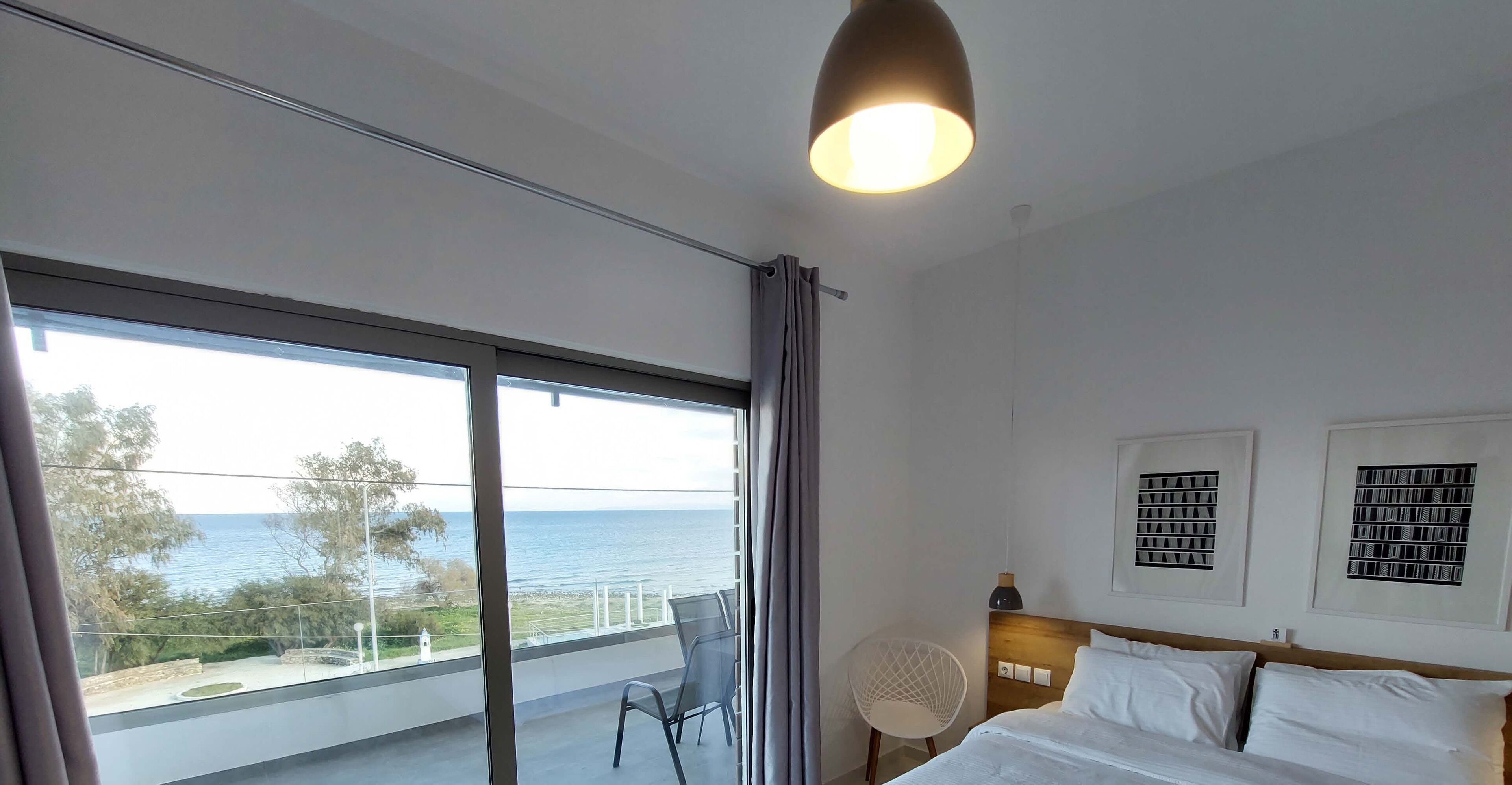 A hotel room with a large window offering a view of the sea, a bed with white linens, a modern chair, and a ceiling light.