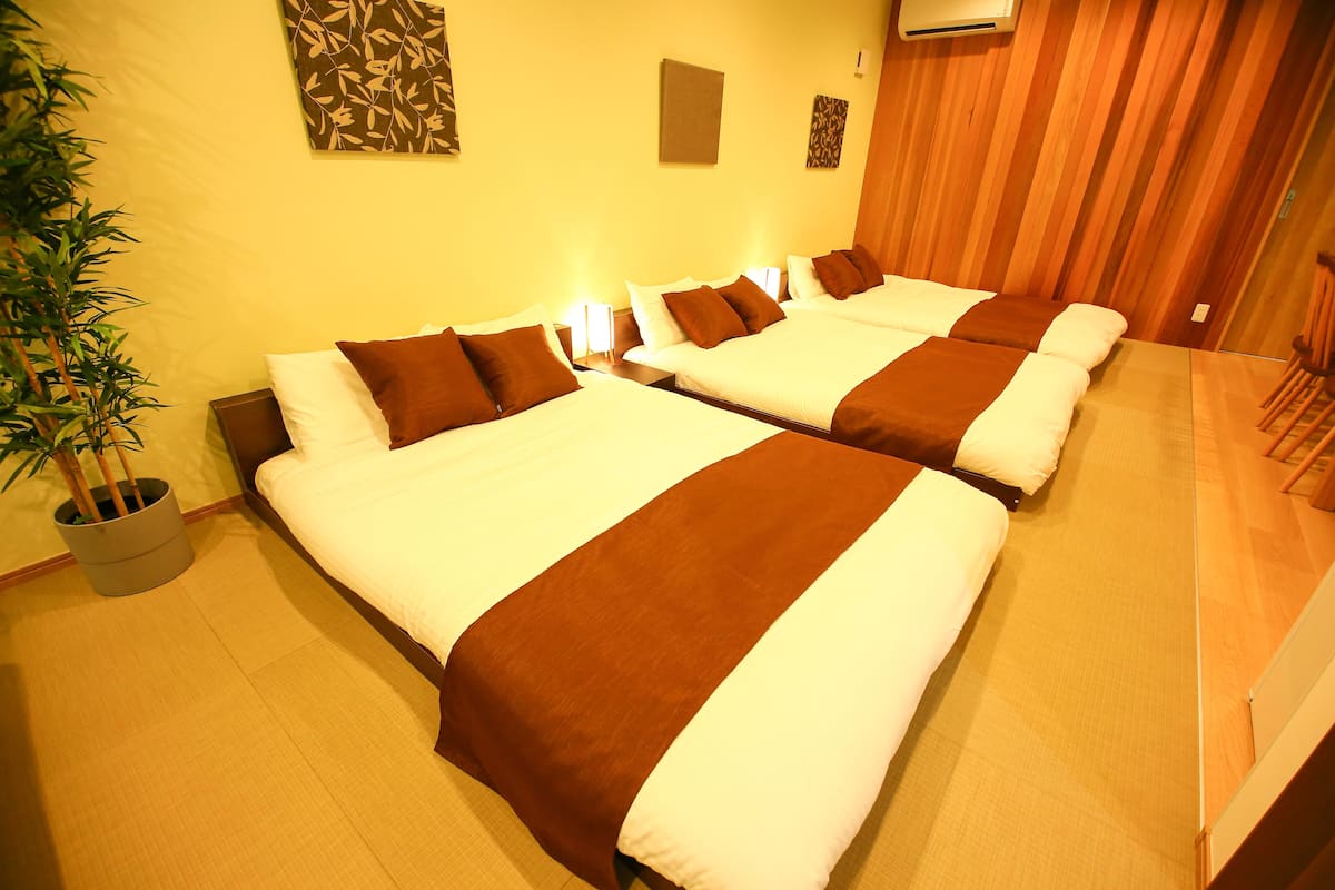 A hotel room with four beds arranged in a row, each with white and brown bedding. There is a potted plant on the left side of the room.