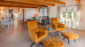 Un'ampia zona giorno con soffitto in legno, un divano grigio, due poltrone color senape, un tavolino rotondo e una finestra con tende.