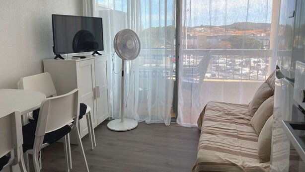 A modern hotel room with a large window, a flat-screen TV, a standing fan, a sofa, and a dining area with chairs.