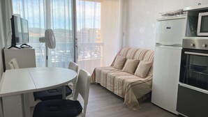 A compact living space with a dining table, chairs, a sofa, a TV, a fan, a refrigerator, and an oven.