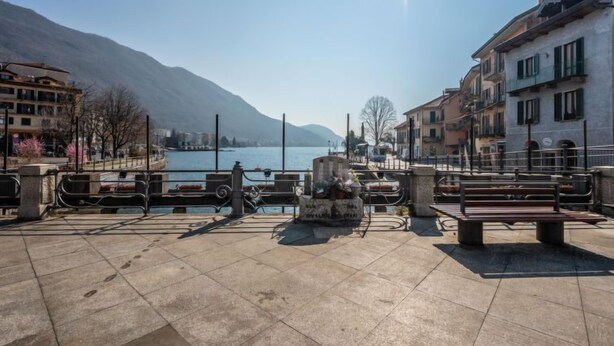 Eine Uferpromenade mit Bänken, einem Steinmonument und Gebäuden am See entlang.