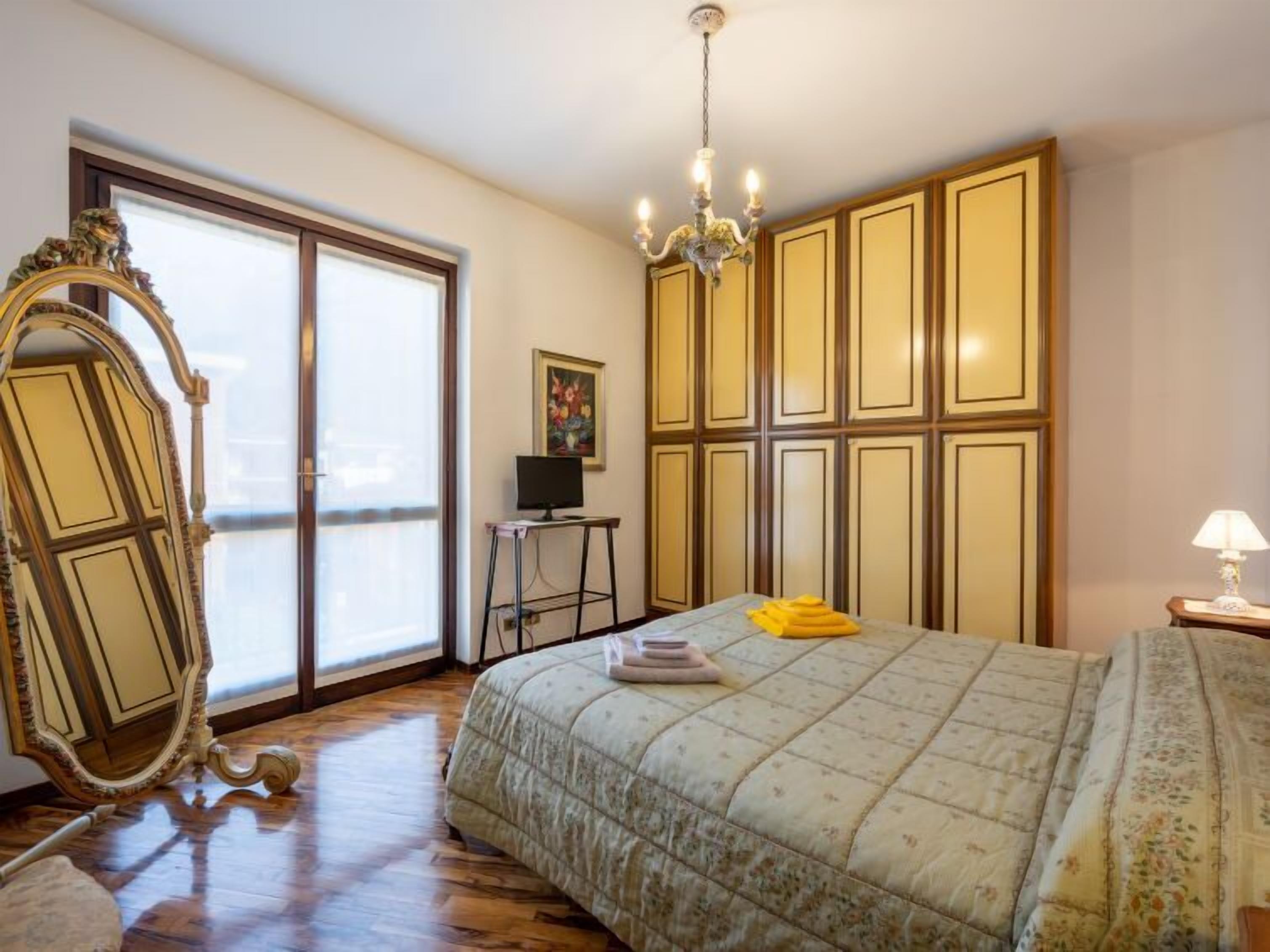 A bedroom with a large bed, a chandelier, a desk with a computer, a large mirror, and a view through a sliding door.