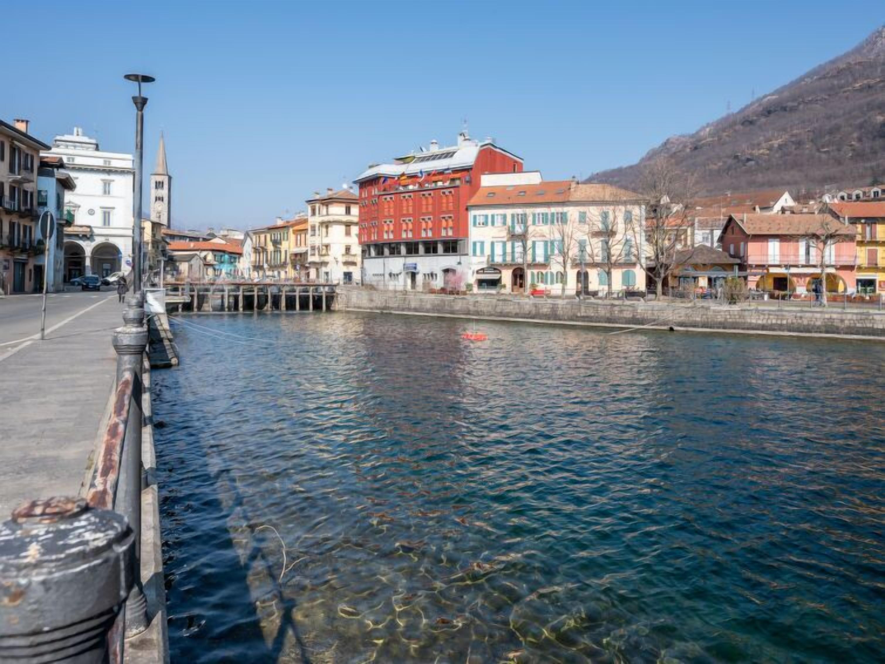 A waterfront promenade with buildings along the riverbank, a clear sky, and a mountain in the background.