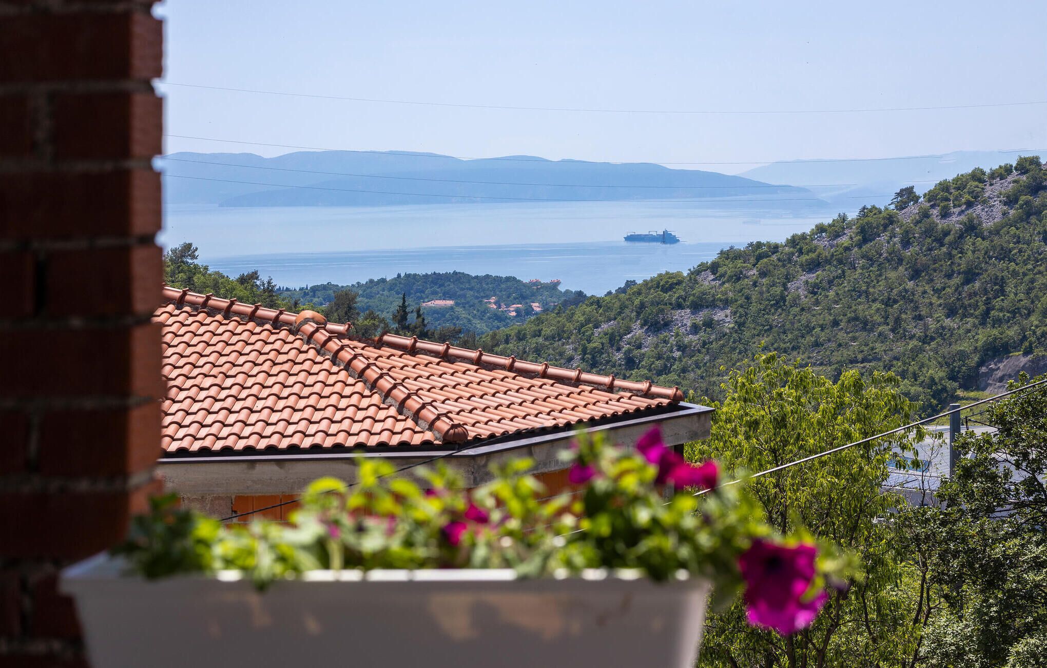 Blick auf die Küste mit einem Ziegeldach, einem Blumenkasten mit Blumen und einem Schiff in der Ferne auf dem Wasser.