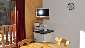 Une pièce avec un plafond en bois, un téléviseur fixé au mur, une petite table avec des chaises, une fenêtre avec des rideaux et une horloge au mur.