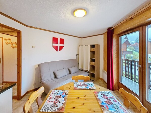 A cozy room with a sofa, a wooden dining table with placemats, a red shield decoration, and a window with a view of houses.