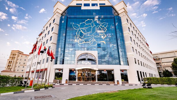 A modern hotel with a large glass facade, a central entrance, and multiple flags. - Peony Hotel (Dubai)