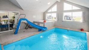 An indoor swimming pool with a blue slide, a small table with a towel, and a window with a view of houses. - Stunning home in Samsø with sauna (Samsø)