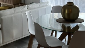 A modern living room with a glass dining table, white chairs, a sofa, and a television on a wooden stand.