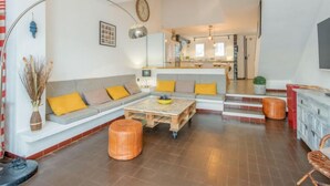 Uma sala de estar moderna com sofá, banquinho e mesa de centro. Uma cozinha é visível ao fundo.