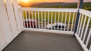 Un balcón con barandilla blanca, suelo de madera y vista a un paisaje montañoso con coches estacionados abajo, bajo un atardecer.