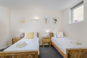 A room with two wooden beds, each with a nightstand and a lamp. There are wall-mounted light fixtures and a window with blinds.