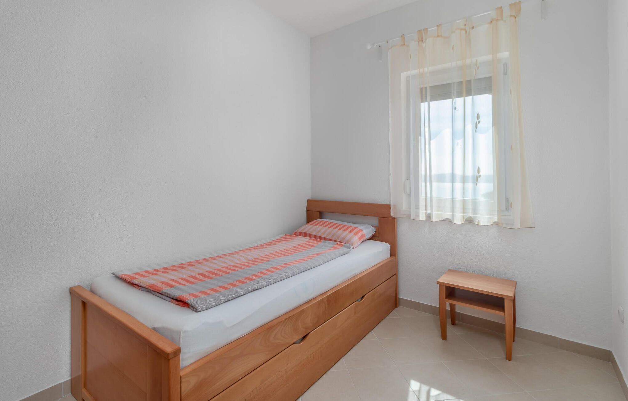 Een eenpersoonsbed met houten frame en een klein nachtkastje in een kamer met eenvoudige wanden en een raam met dunne gordijnen.