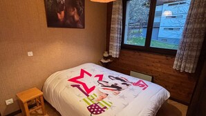 A bedroom with a wooden floor, a bed with a white bedspread featuring a dog and star design, a small wooden stool, a window with curtains, and a wall-mounted lamp. - 2-room apartment near slopes with balcony (La Chapelle-d'Abondance)