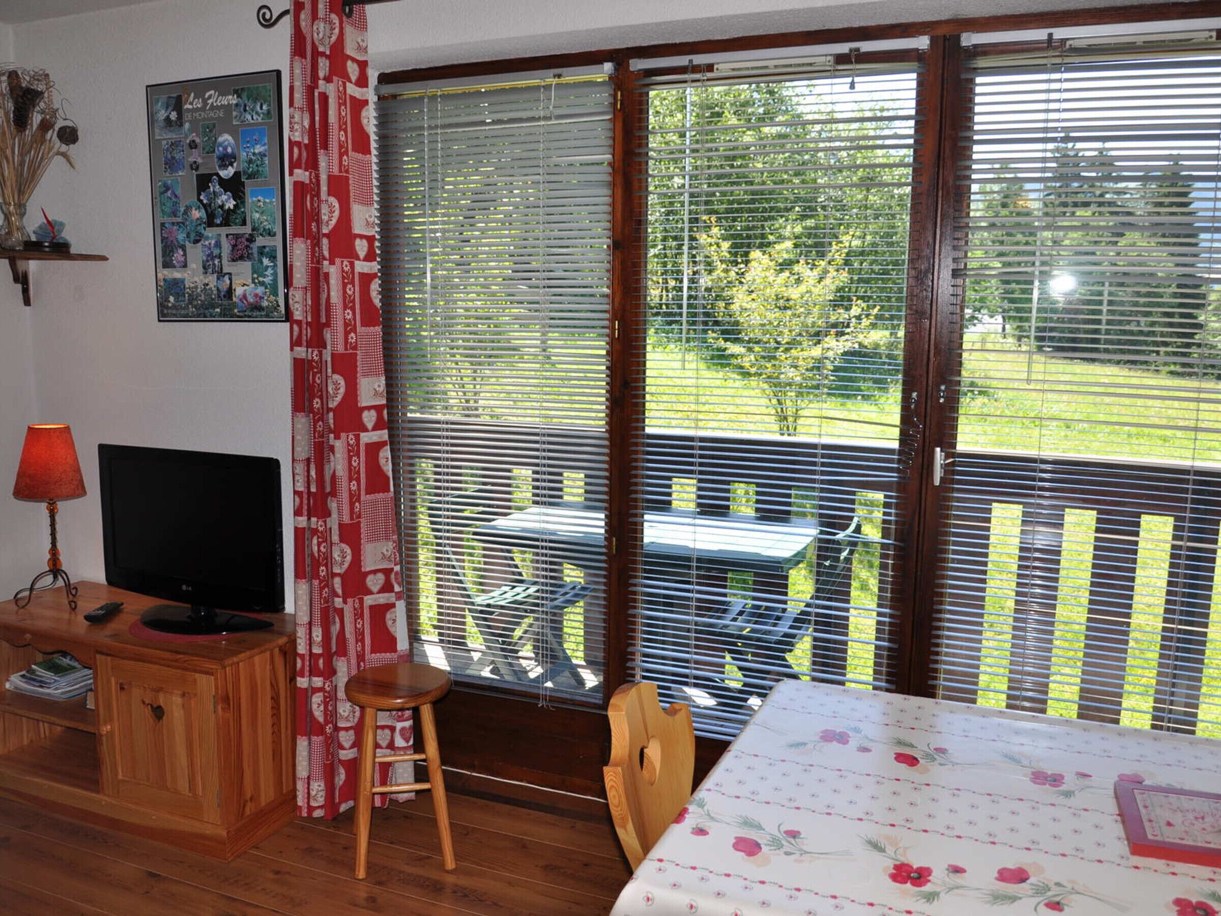 Una habitación con escritorio de madera, televisión, cortina roja y vista a un jardín a través de amplias ventanas.