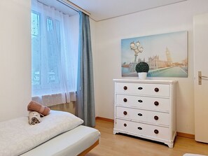 Quarto com cama, cômoda branca, janela com cortinas leves e pintura de paisagem urbana na parede.