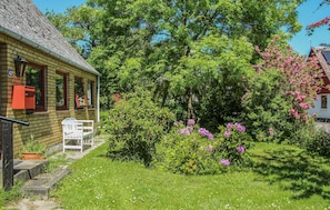 Ein Haus mit Ziegelaußenwand, einer kleinen Veranda und einem Garten mit blühenden Blumen und einem großen Baum.