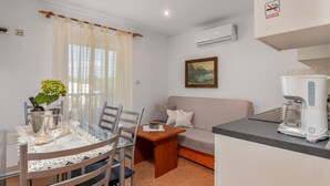 A modern kitchen with a dining area, featuring a coffee maker, a painting, and a window with sheer curtains.