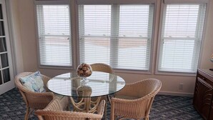 Een kamer met een ronde tafel met glazen blad, omringd door rieten stoelen, grote ramen met zonwering en een deur met ruit.