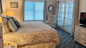 Een slaapkamer met een bed, een stoel, een televisie, een raam met gordijnen en een wanddecoratie.
