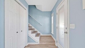 Un couloir avec un escalier, des portes blanches et une fenêtre avec des stores.