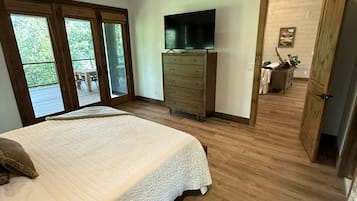 Une chambre dotée d’un grand lit, d’un plancher en bois, d’une commode, d’un téléviseur et d’une porte-fenêtre coulissante donnant sur un espace extérieur.