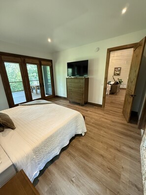 Un dormitorio con una cama grande, suelo de madera, un aparador, un televisor y un ventanal corredero que da a un espacio exterior.