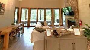 Ein geräumiges Zimmer mit hoher Decke, Holzbalken, einem Kamin aus Stein, einem Fernseher und einem Essbereich mit Holztisch und Stühlen.