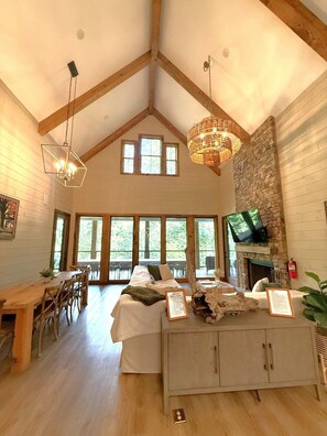 A spacious room with a high ceiling, wooden beams, a stone fireplace, a television, and a dining area with a wooden table and chairs. - Luxury river home on the Chattahoochee River (Demorest)