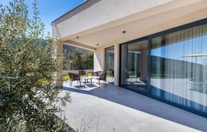 Enjoy dining or socializing in this outdoor patio area with a table and chairs, surrounded by lush vegetation and a glass door leading to an adjacent room.