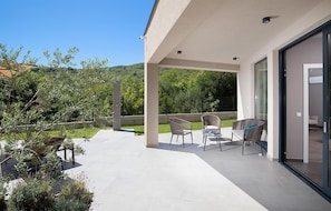 A covered outdoor patio with a table and chairs, a glass door leading to a room, and a view of a green landscape.