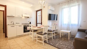 A modern kitchen with white cabinets, a gas stove, and a built-in oven. There is a dining area with white chairs and a table, a sofa, and a TV mounted on the wall. - Cozy apartment in Quartu Sant'Elena with WiFi (Quartu Sant'Elena)