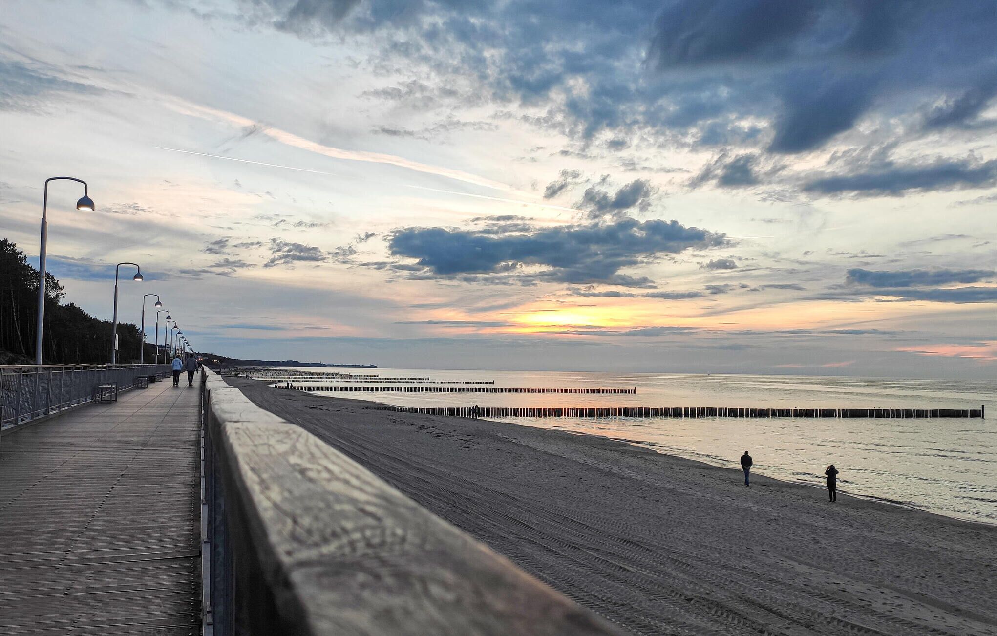 Un paseo marítimo costero con una pasarela de madera y farolas, que conduce a una playa arenosa con un muelle que se extiende hacia el agua.