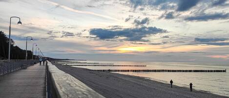 Un paseo marítimo costero con una pasarela de madera y farolas, que conduce a una playa arenosa con un muelle que se extiende hacia el agua.
