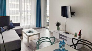 A modern living room with a sofa, a coffee table, a TV mounted on the wall, and a dining area with chairs and a table. - 1 bedroom cozy apartment in Koszalin (Koszalin)