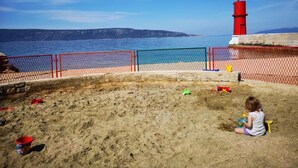 A sandy beach area with a red and blue fence, a lighthouse, and a child playing. - Naomi's Sunshine (Cres)