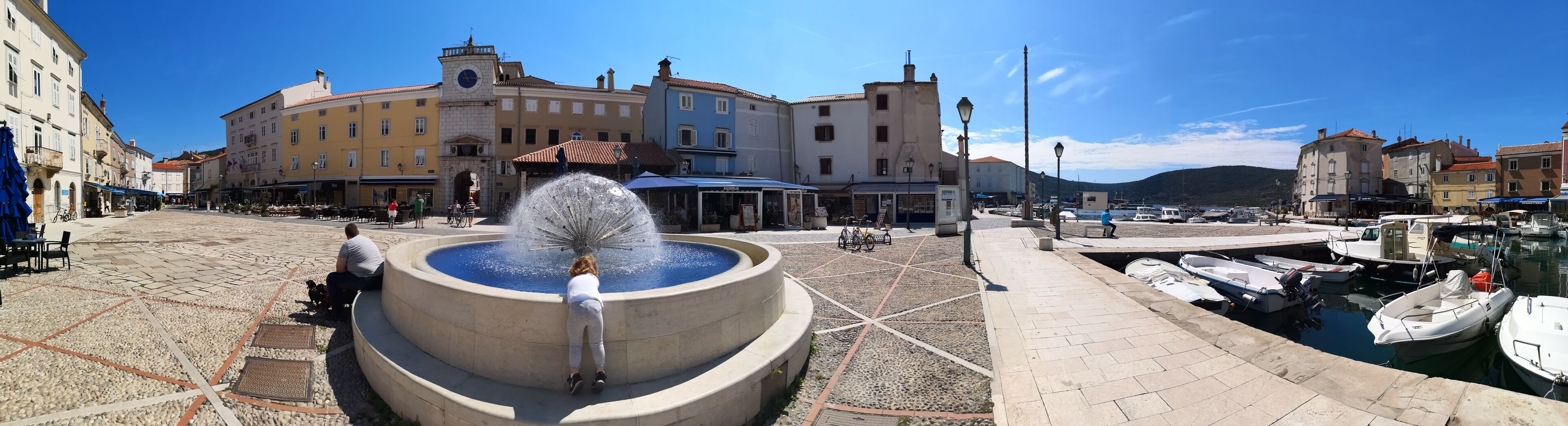 A public square with a fountain, a clock tower, and a marina with boats.