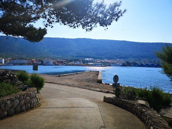 Un paseo marítimo a la costa que conduce a un puerto con barcos, edificios y montañas al fondo.