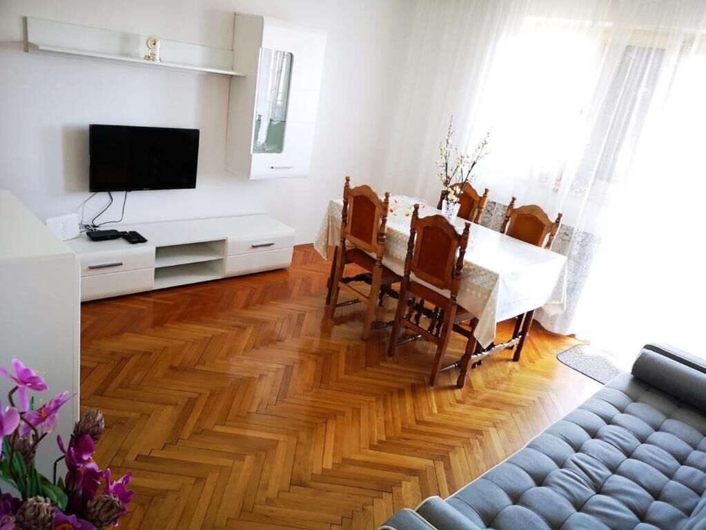 Un salon moderne avec un parquet en bois disposé en motif chevron, un canapé gris, une table à manger avec des chaises, un téléviseur à écran plat fixé au mur et une fenêtre agrémentée de rideaux légers.