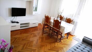 Un salon moderne avec un parquet en bois disposé en motif chevron, un canapé gris, une table à manger avec des chaises, un téléviseur à écran plat fixé au mur et une fenêtre agrémentée de rideaux légers.