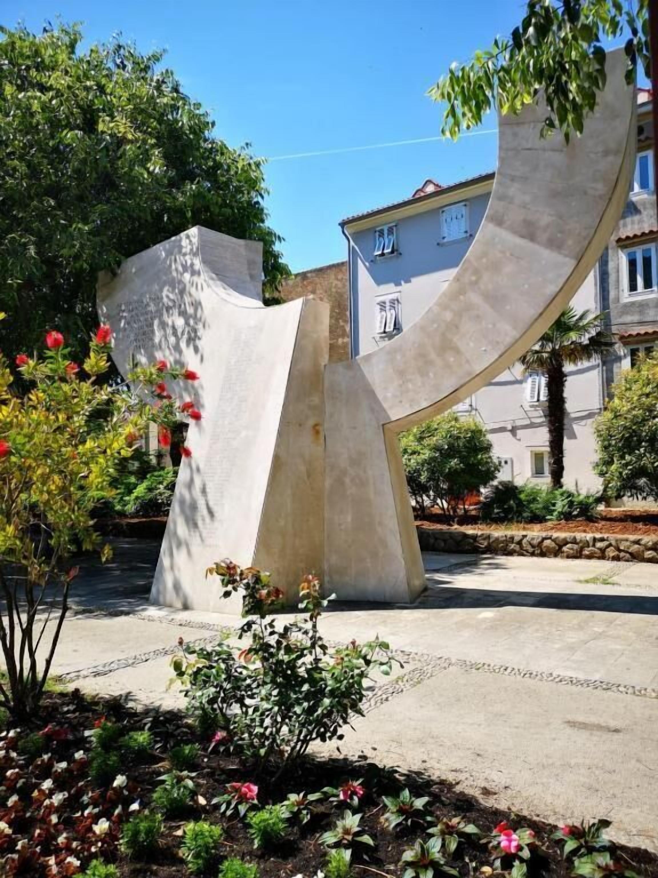 A modern sculpture in a garden area with a paved walkway and surrounding greenery.