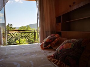Quarto com cama, almofadas florais, vista para áreas verdes e colinas, e varanda com guarda-corpo.