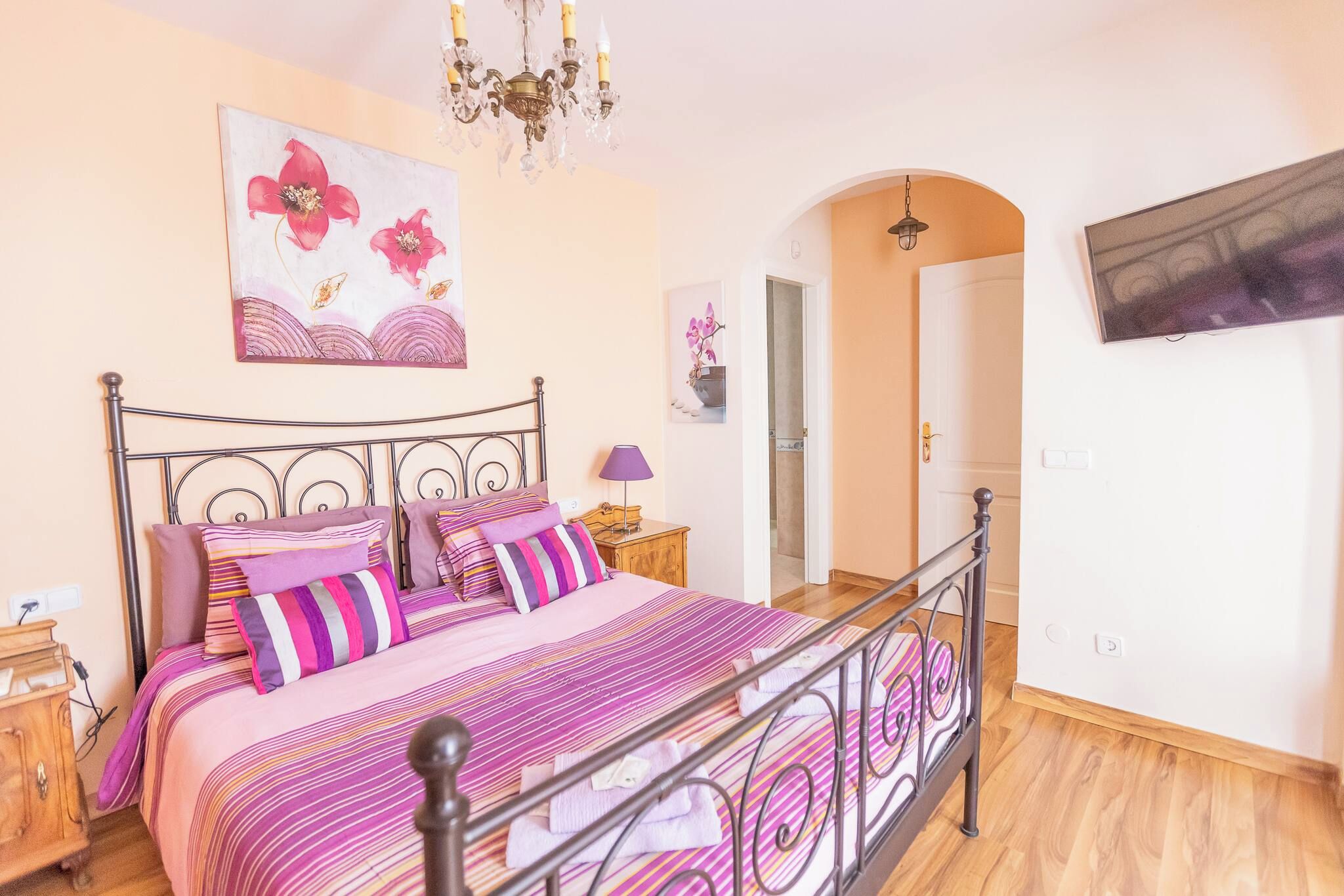 A bedroom with a bed, a nightstand, a lamp, a painting, and a door leading to another room.