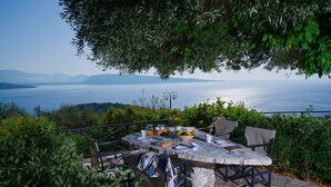 An outdoor dining area with a stone table and chairs, surrounded by greenery and overlooking a body of water with mountains in the distance. - Villa Vasso Two-Bedroom Sea View Residence near Kerasia Beach (Αγνίτσινη)