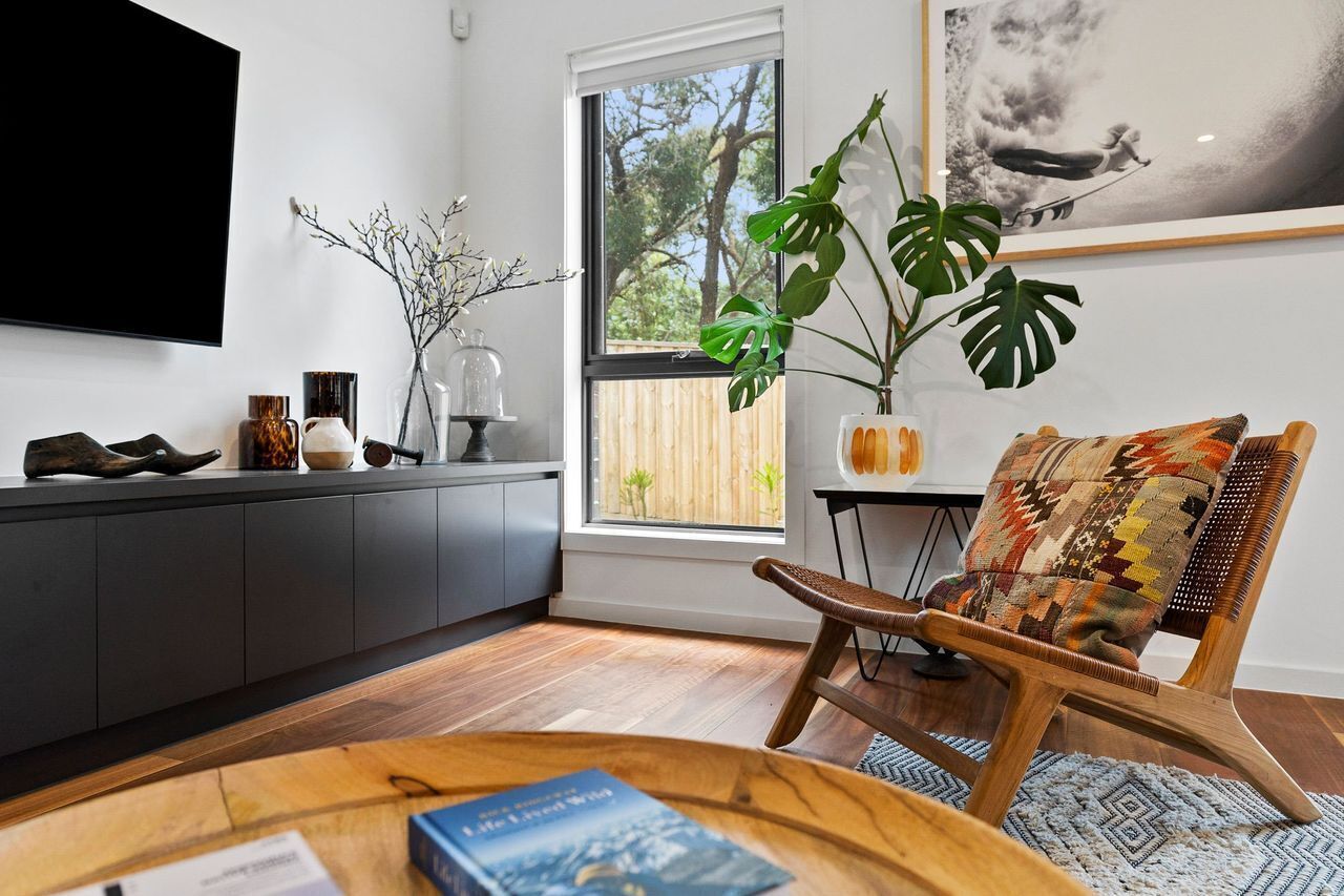 Una sala de estar moderna con una mesa de centro de madera, un sillón estampado, un ventanal con vistas a árboles y un televisor montado.
