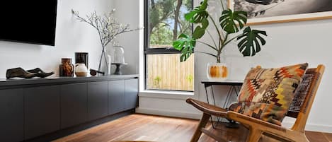 Una sala de estar moderna con una mesa de centro de madera, un sillón estampado, un ventanal con vistas a árboles y un televisor montado.