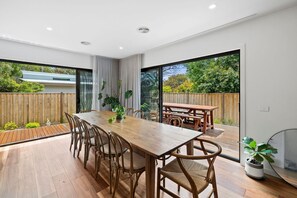 A modern dining area with a wooden table and chairs, large windows, and a view of the outdoors. - McMillan Escape (Anglesea)